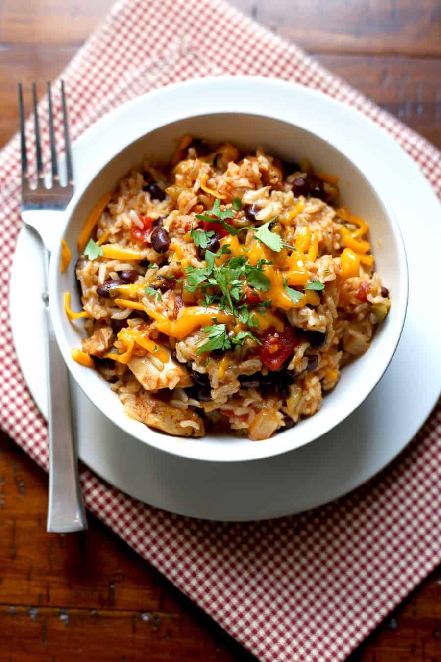 Chicken, Black Bean, and Rice Burrito Bowls in white bowl topped with cheese and chopped cilantro. A fork sits beside the bowl.