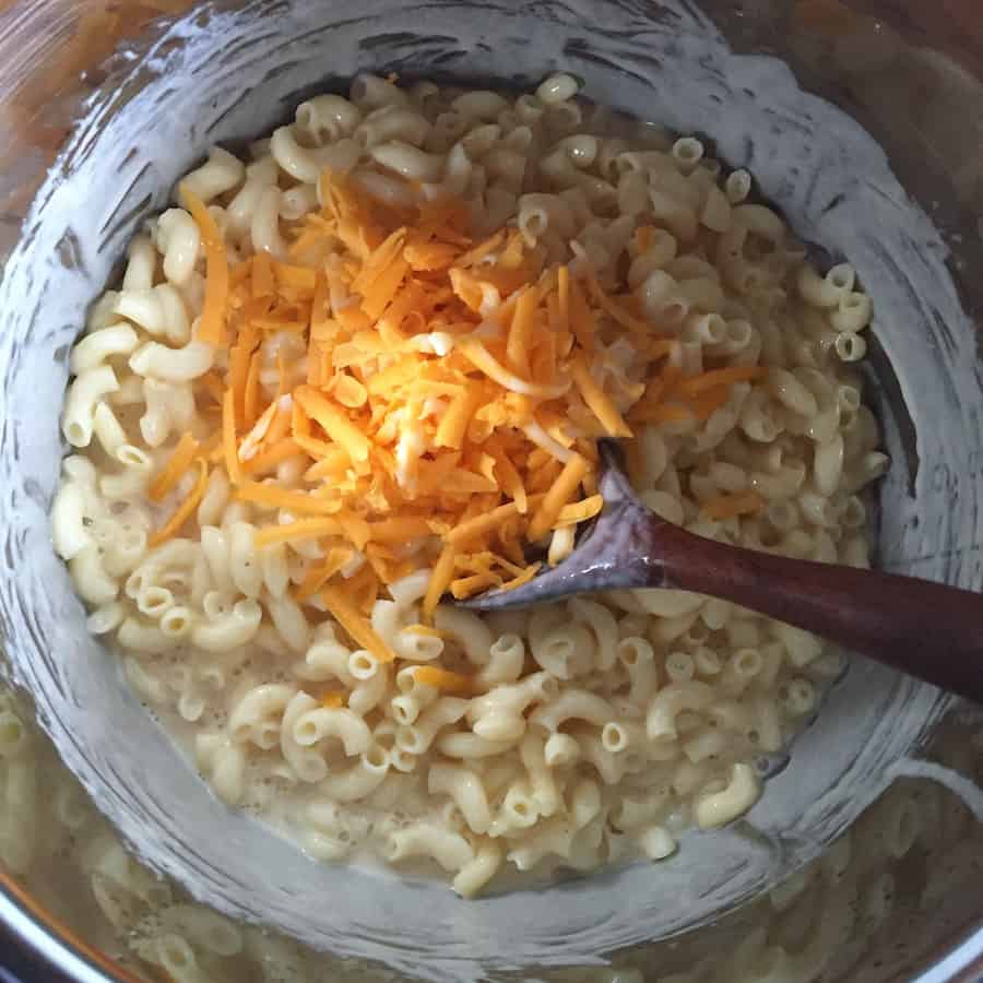 Cooked macaroni in a pressure cooker pot with shredded cheese. A wooden spoon stirs the mixture.