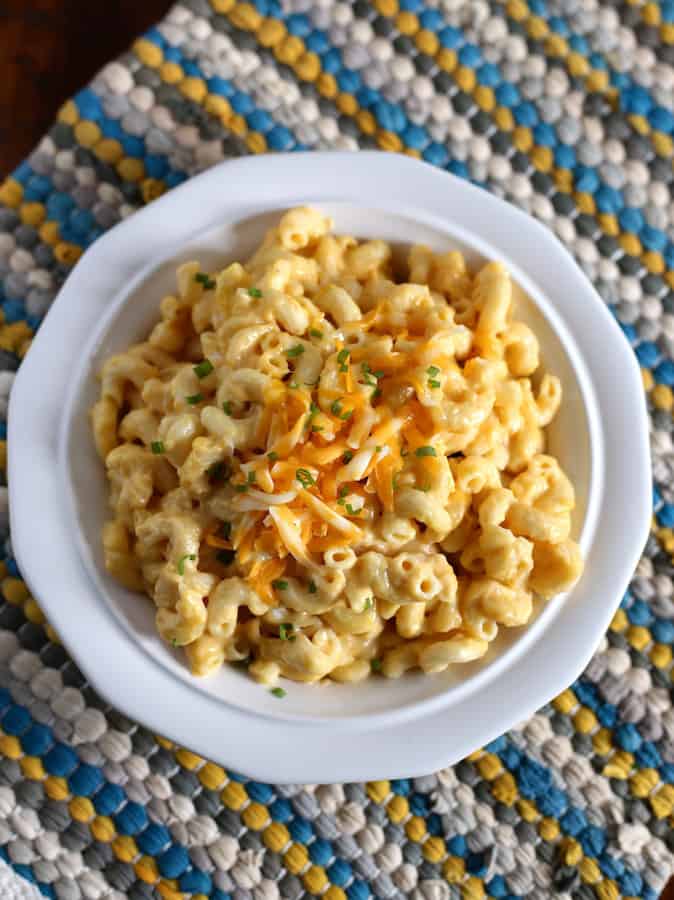 Cooked macaroni and cheese in white bowl. Topped with grated cheese and scallions. The bowl is a blue and yellow woven placemat.