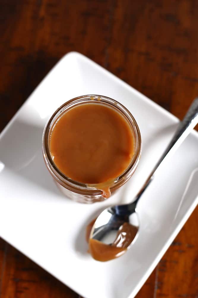 Caramel sauce in a jelly jar on a white plate with a spoon sitting next to it.