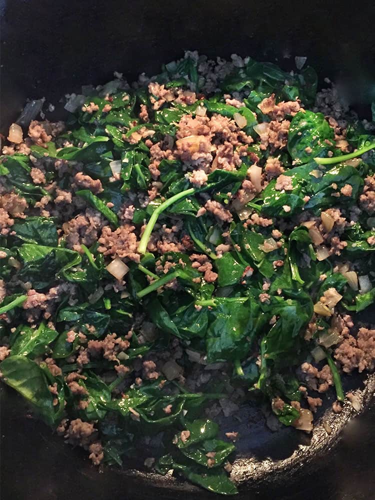 Cooked, wilted spinach stirred together with cooked sausage and onions in a black cast iron pan.