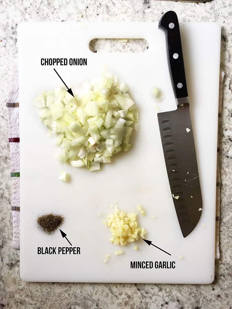 A white cutting board with a chopped onion, cracked black pepper, and minced garlic. To the right sits a Chef's knife. 