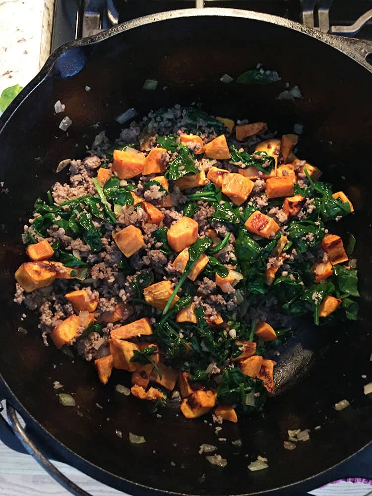 Cubes of roasted sweet potatoes in a black cast iron pan with cooked spinach, sausage, and onions.