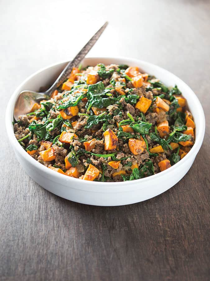 Cooked sweet potato hash with spinach and sausage in a white serving dish. A serving spoon is on left side of the dish.