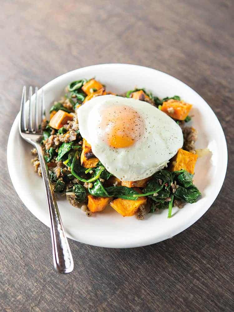 A cooked eggs sits on a plate of sweet potato hash with spinach and sausage. A fork is on the left of the plate.