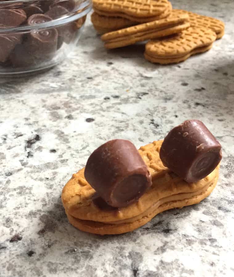 A Nutter Butter cookie on a counter with two Rolo candies attached to it. In the background: a pile of Nutter Butter cookies and a bowl of Rolo candies.