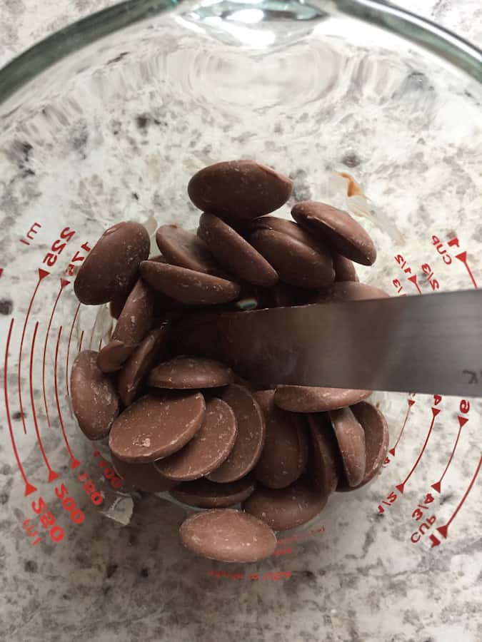 Chocolate melting wafers in a measuring cup. A metal spatula stirs the wafers.