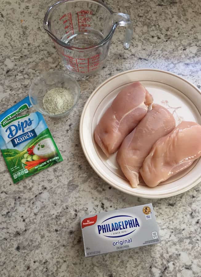 Ingredients for Pressure Cooker Crack Chicken Recipe. Raw chicken on plate. Block of cream cheese. Packet of ranch dressing. Water in measuring cup.