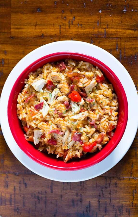 Beer can chicken with rice in a red bowl. Pieces of cooked chicken and bacon are on top of the cooked rice.