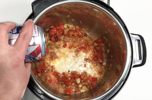 Can of beer being poured into a pressure cooker pot with cooked onions, red bell peppers, and onions.