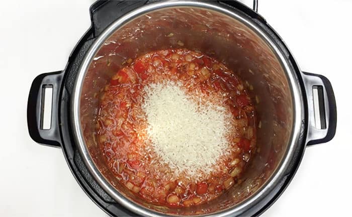 Uncooked rice sitting in a pressure cooker pot filled with beer, cooked red peppers, onions, and garlic.