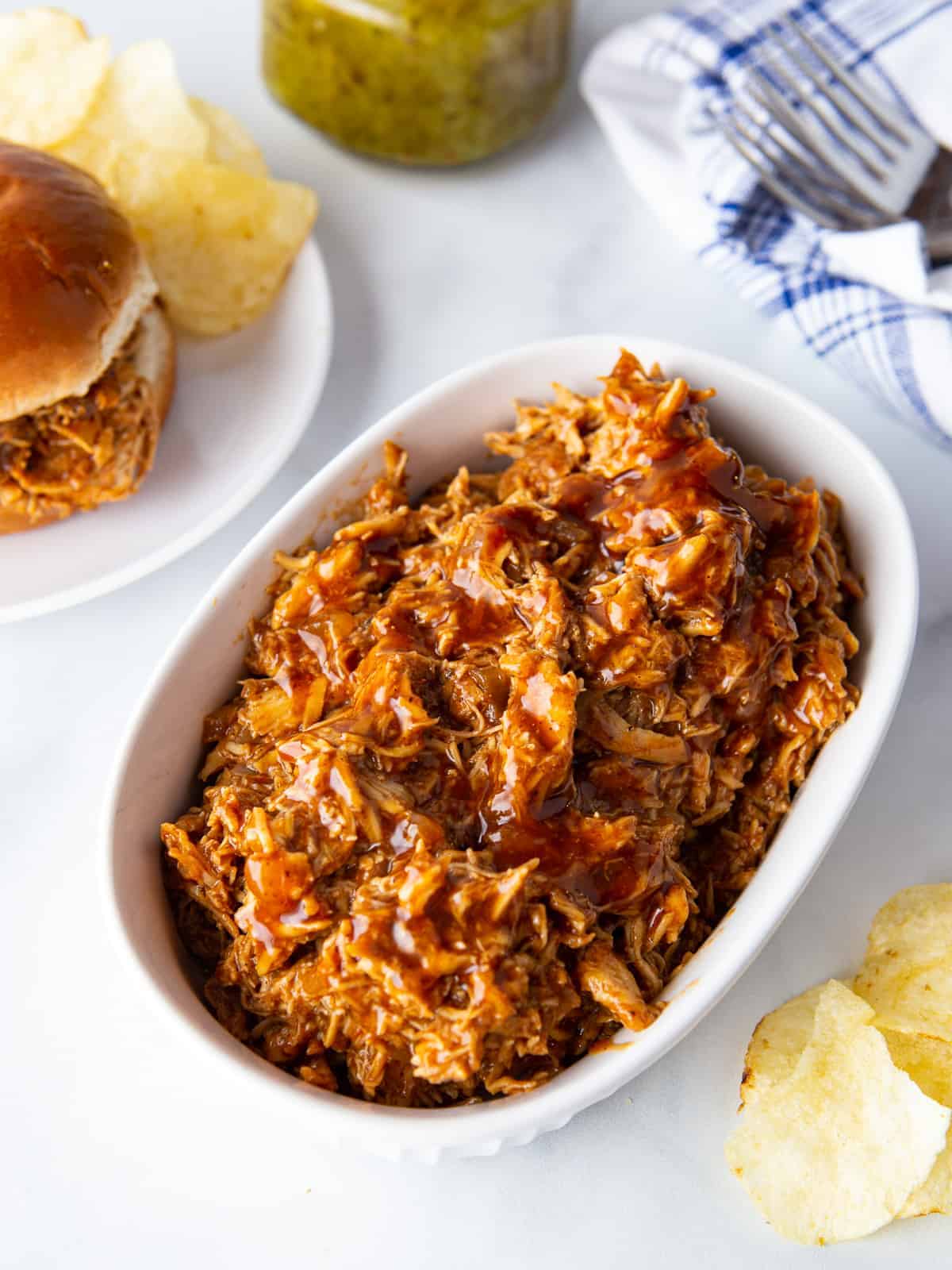 Serving dish with barbecue pulled chicken. On the side is a pulled chicken sandwich on a plate with potato chip.