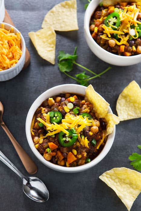 Two bowls with chili topped with grated cheese and jalapeño slices. Tortilla chips and a bowl of shredded cheese sit next to the bowls.