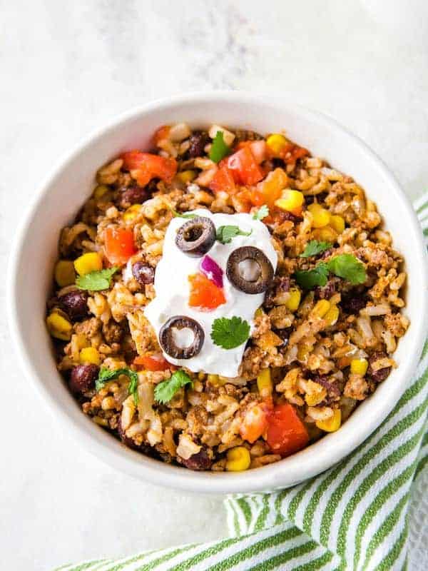 White bowl filled with cooked rice, black beans, corn, and ground beef. Topped with sour cream and salsa.