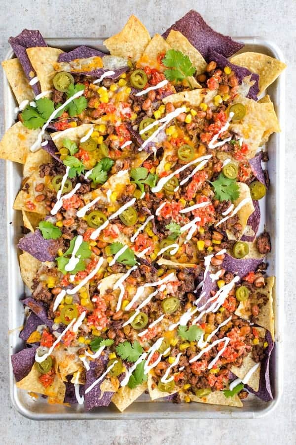 Sheet Pan with Baked Nachos. Topped with Cooked Ground Beef, Corn, Salsa, Drizzled Sour Cream, Beans, and Chopped Cilantro