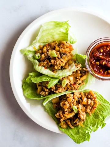 Three chicken lettuce wraps on a plate. A small bowl of chili sauce sits alongside the wraps.