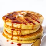 Three cornmeal pancakes stacked on a plate with maple syrup and butter.