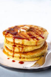 Three cornmeal pancakes stacked on a plate with maple syrup and butter.