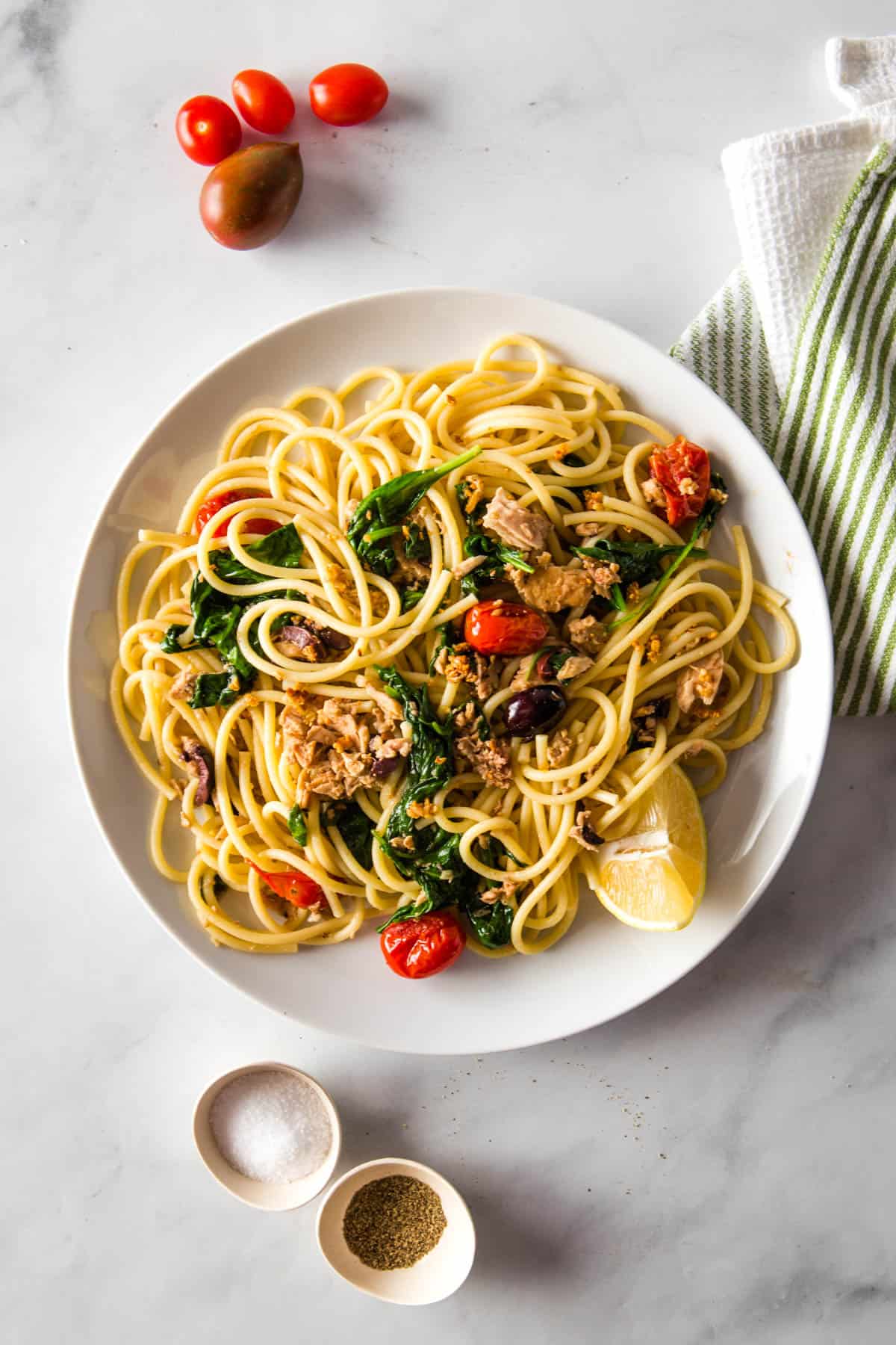 Bowl of pasta with tuna, spinach, tomatoes, and olives. A lemon sits alongside the pasta.