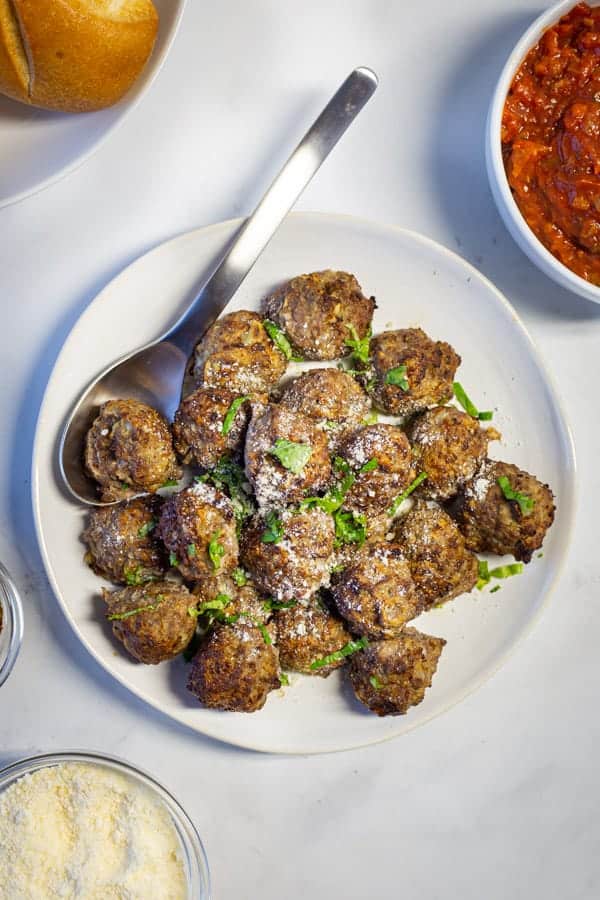Plate of air fryer meatballs. Topped with chopped basil and sprinkled with cheese. A bowl of sauce and a bowl of grated cheese are alongside the meatballs.