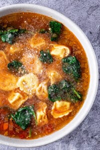 Bowl of tortellini soup with spinach topped with grated parmesan cheese.