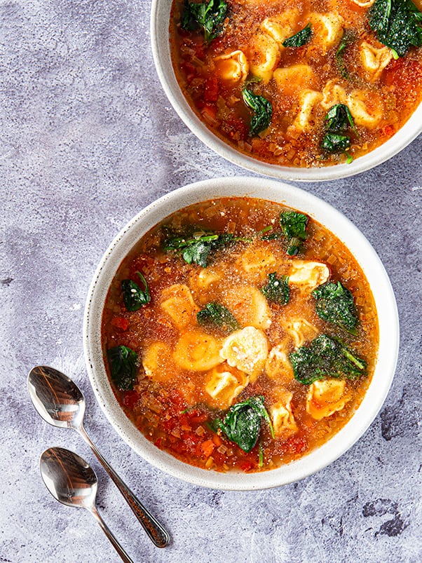 Two bowls of tortellini soup with spinach and topped with grated parmesan sit on a gray marble countertop.