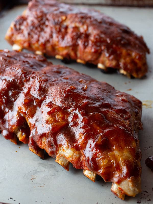 Two racks of ribs on a baking sheet. The ribs are covered with BBQ sauce.