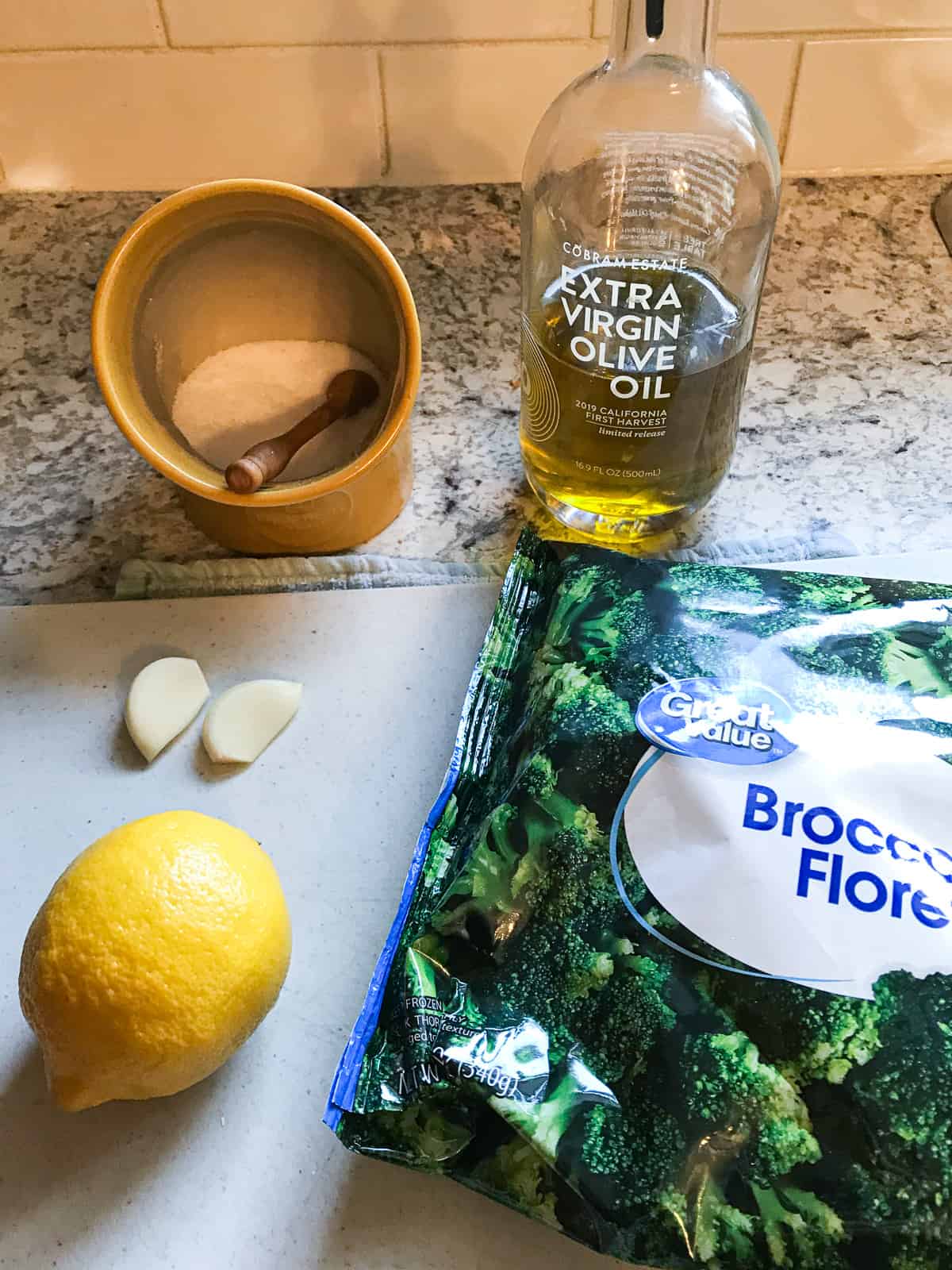 Ingredients for cooking frozen broccoli. Salt, olive oil, bag of broccoli florets, lemon, and two cloves of garlic.