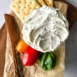 Bowl of herb and garlic cream cheese on a cutting board. Crackers and grape tomatoes are served alongside.