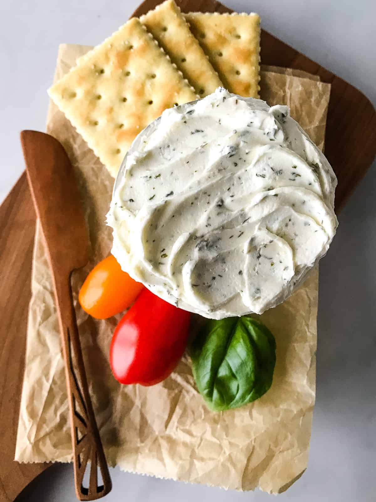 Bowl of herb and garlic cream cheese on a cutting board. Crackers and grape tomatoes are served alongside.