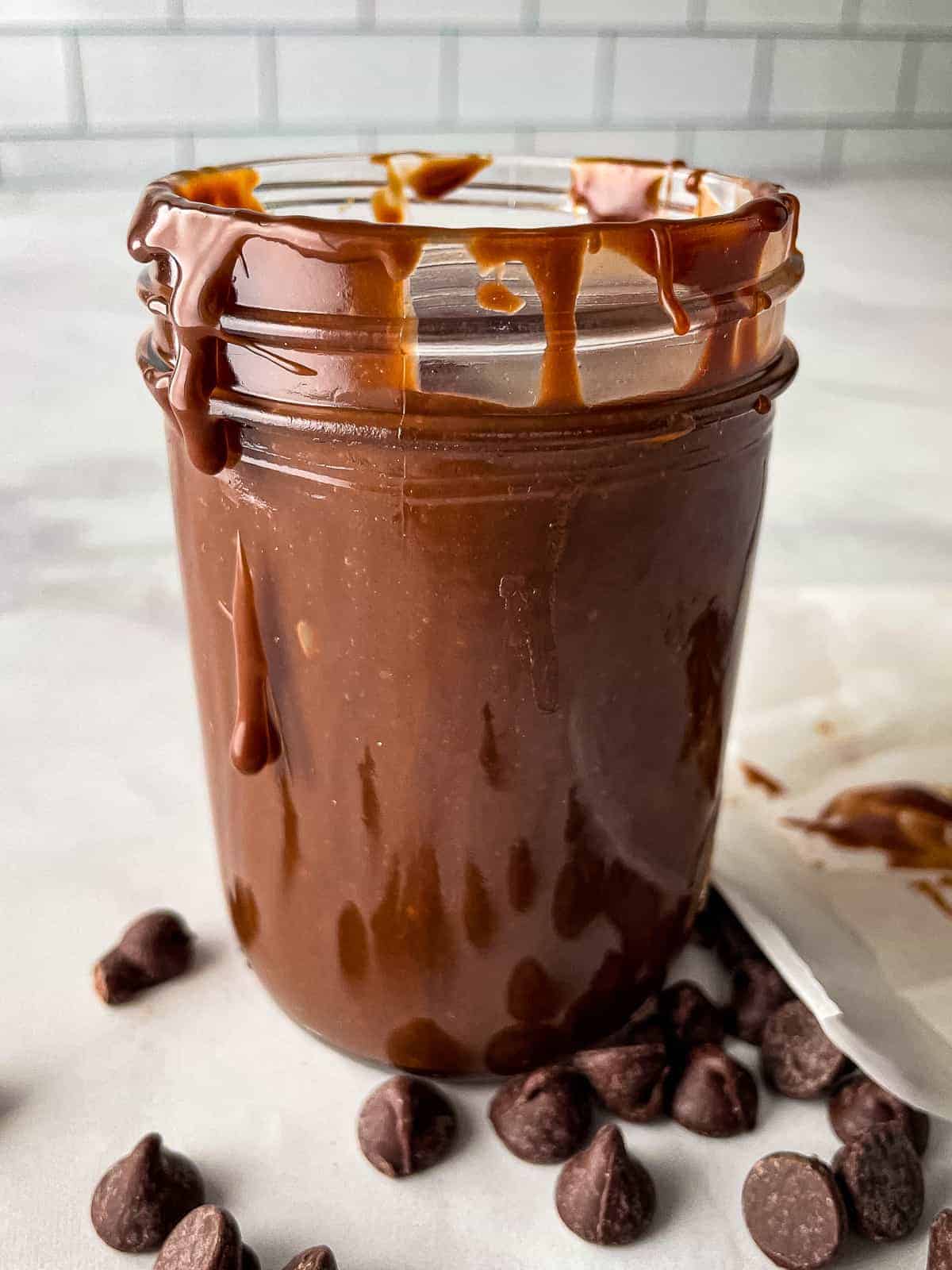 Hot fudge sauce in a glass jar with drips down the sides. Chocolate chips are on the counter next to the jar.
