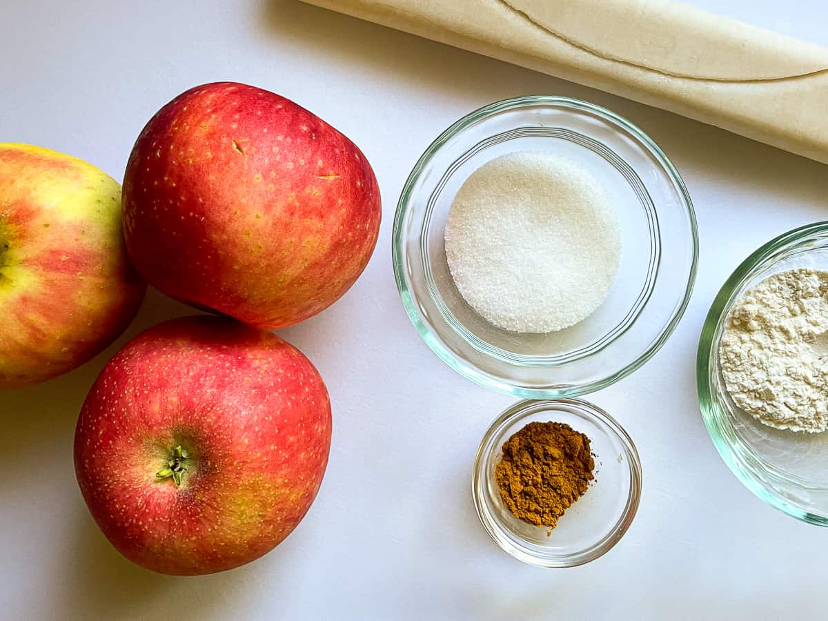 Apples, pie dough, sugar, cinnamon, and flour on a white board.