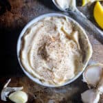 Garlic mayonnaise in a bowl with black pepper on top. Spoon, lemon slices, and garlic cloves off to the side.