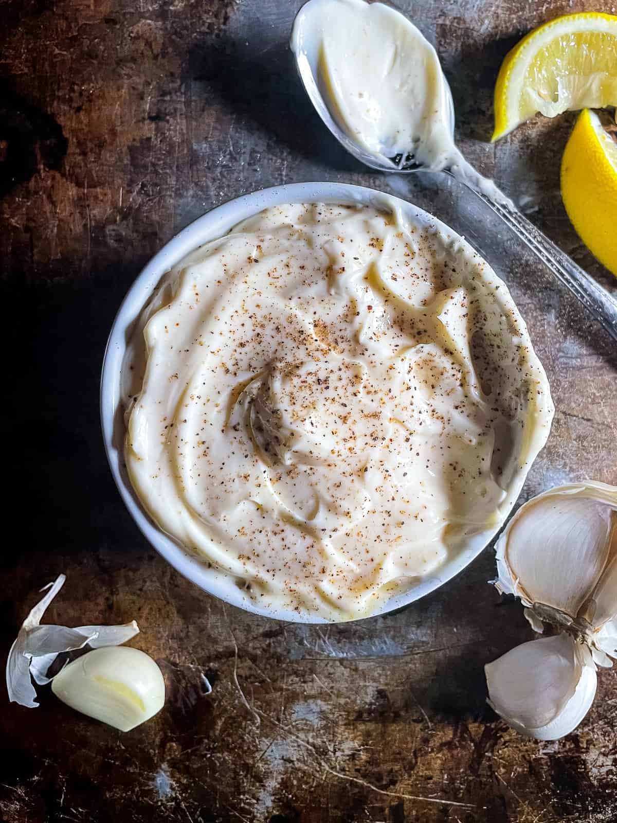 Garlic mayonnaise in a bowl with black pepper on top. Spoon, lemon slices, and garlic cloves off to the side. 