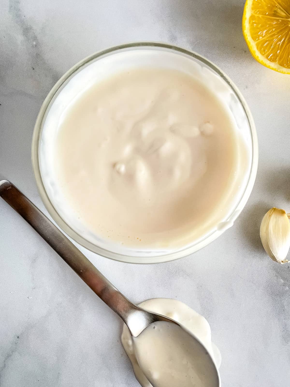 Bowl of garlic mayonnaise. Spoon with mayonnaise. Lemon and garlic cloves off to the side.