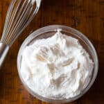 Bowl of whipped cream on a wood table with a whisk sitting next to it.