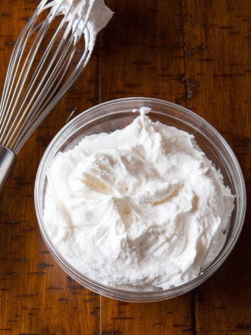 Bowl of whipped cream on a wood table with a whisk sitting next to it.