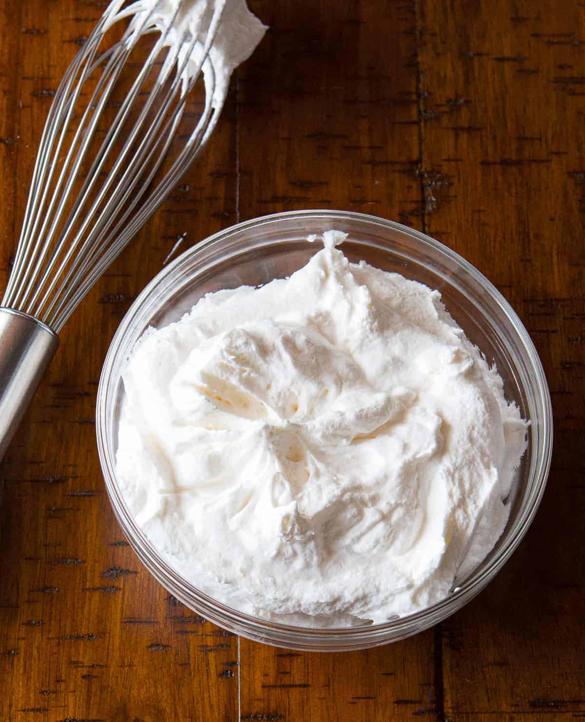 Bowl of whipped cream on a wood table with a whisk sitting next to it.