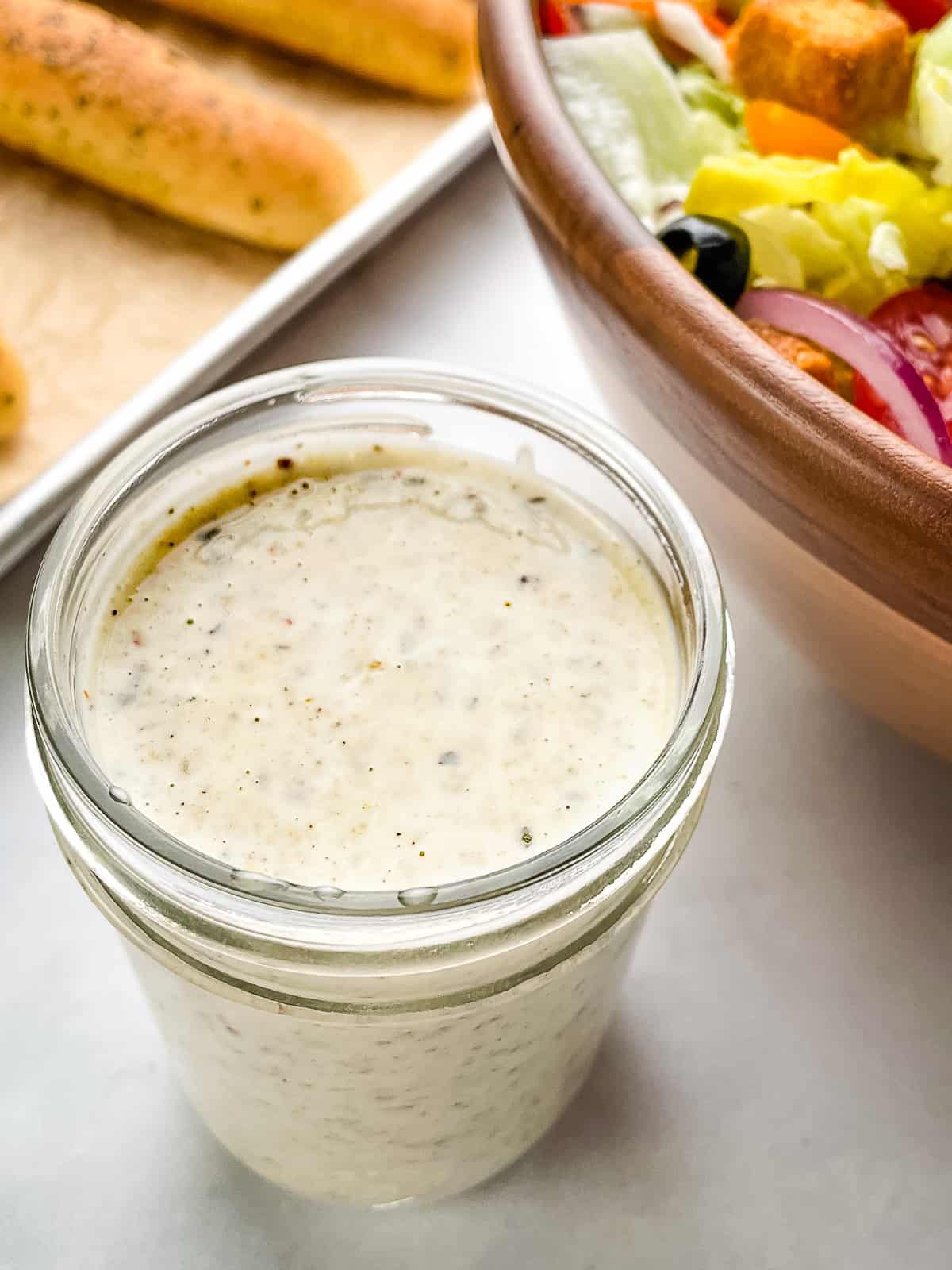 Pint jar filled with creamy Italian dressing. Salad and breadsticks in the background.