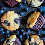 Baked blueberry muffins in a pan. Fresh blueberries are scattered around the pan.