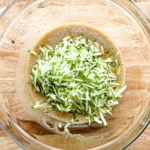 Adding shredded zucchini to egg, oil, sugar mixture in glass bowl.