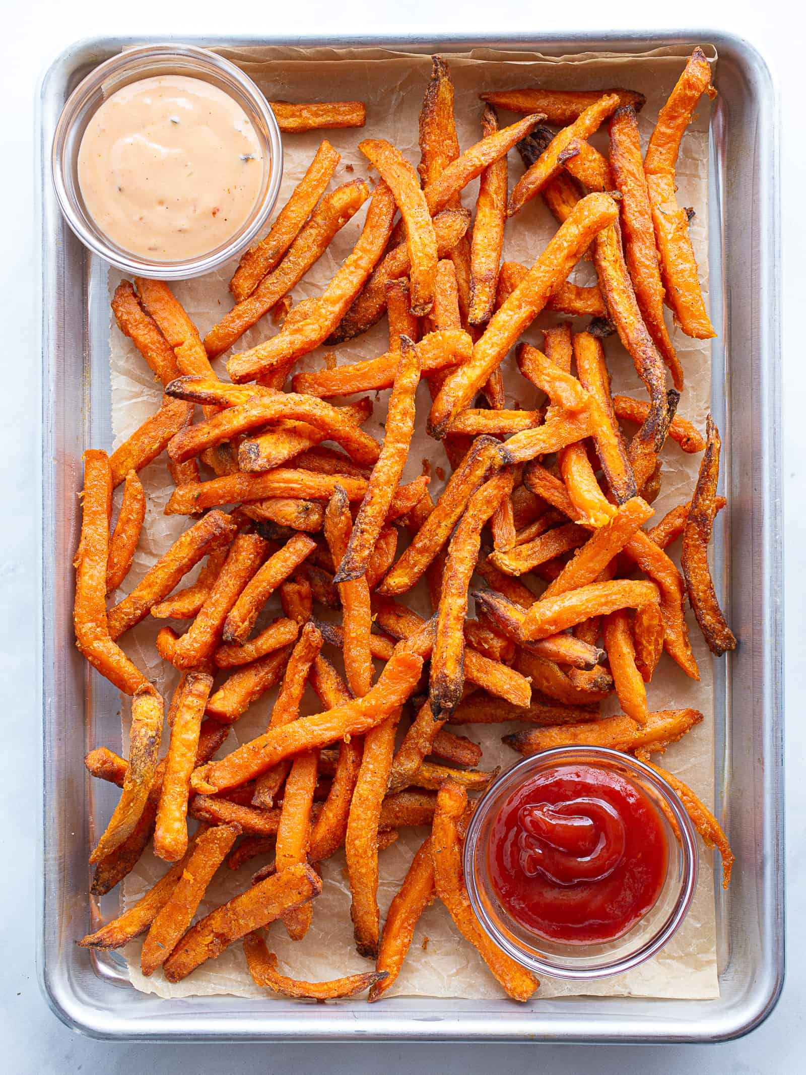 Cooked frozen sweet potato fries on a sheet pan. Spicy mayo in a bowl, upper left corner. Ketchup in bowl, lower right.