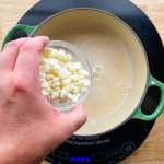 Adding white chocolate chips to a pot of white chocolate sauce.