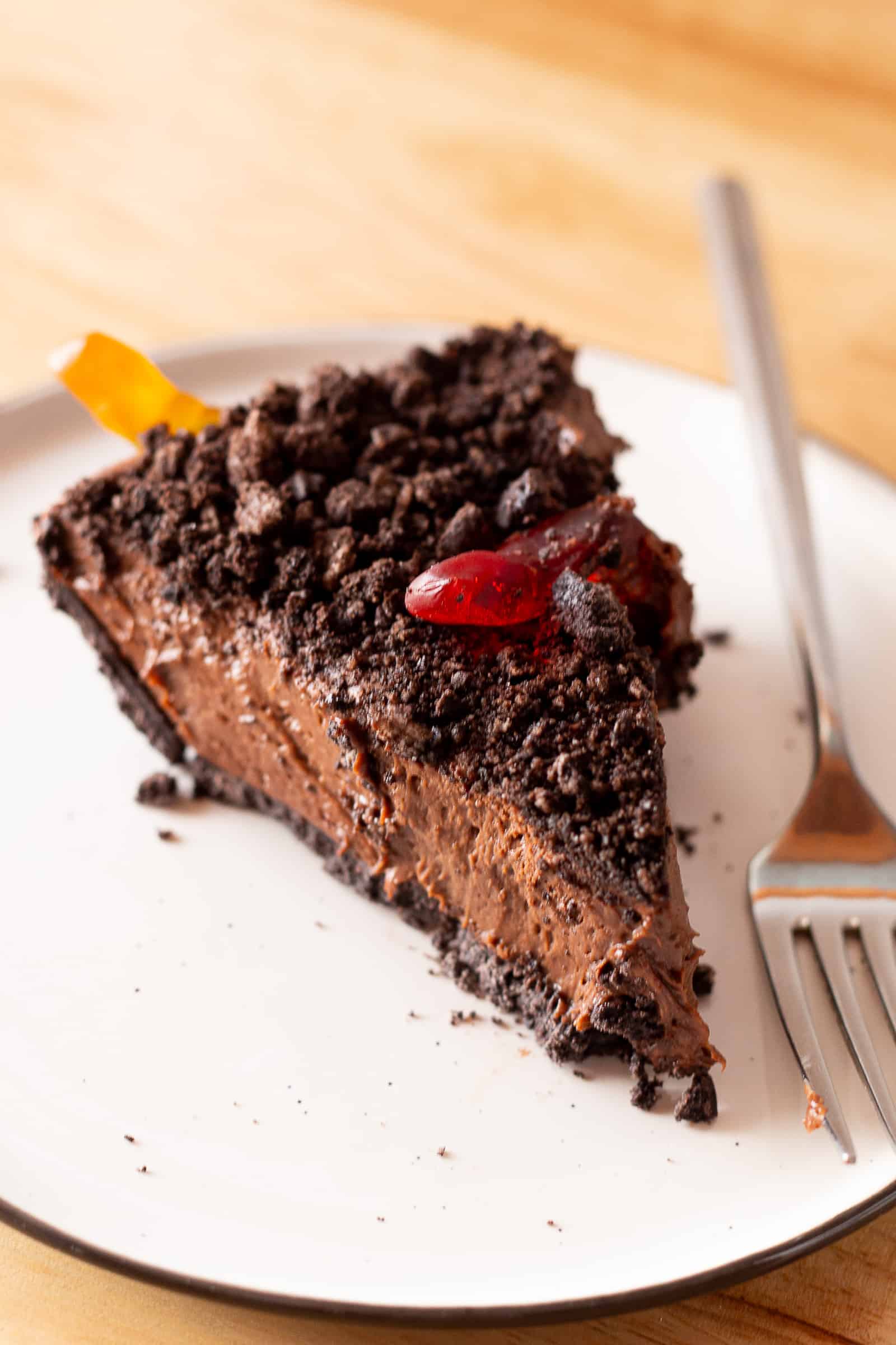 Slice of chocolate dirt pie on a plate. The pie is topped with crushed cookies and two gummy worms.