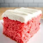 Slice of strawberry cake on a plate. Cake is frosted with cream cheese frosting.