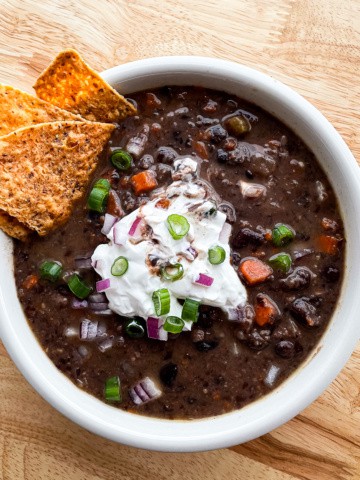 Bowl of black bean soup topped with sour cream, diced red onion, and chopped scallions. A few tortilla chips are on the side of the soup.