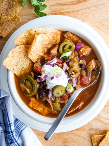 A bowl of chicken tortilla soup topped with sour cream, minced red onions, pickled jalapeños, and tortilla chips.