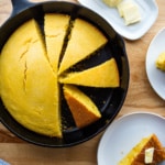 Cornbread sliced in a skillet. Off to the side, a stick of butter on a holder and a slice of cornbread on a plate.