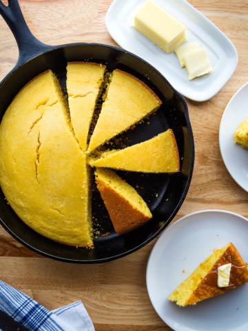 Cornbread sliced in a skillet. Off to the side, a stick of butter on a holder and a slice of cornbread on a plate.