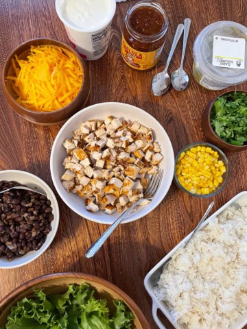 Dinner table set with the makings of chicken burrito bowls. Clockwise from left: shredded Cheddar cheese. Sour cream. Salsa. Guacamole. Chopped cilantro. Corn. White rice. Lettuce. Black beans. Diced grilled chicken.
